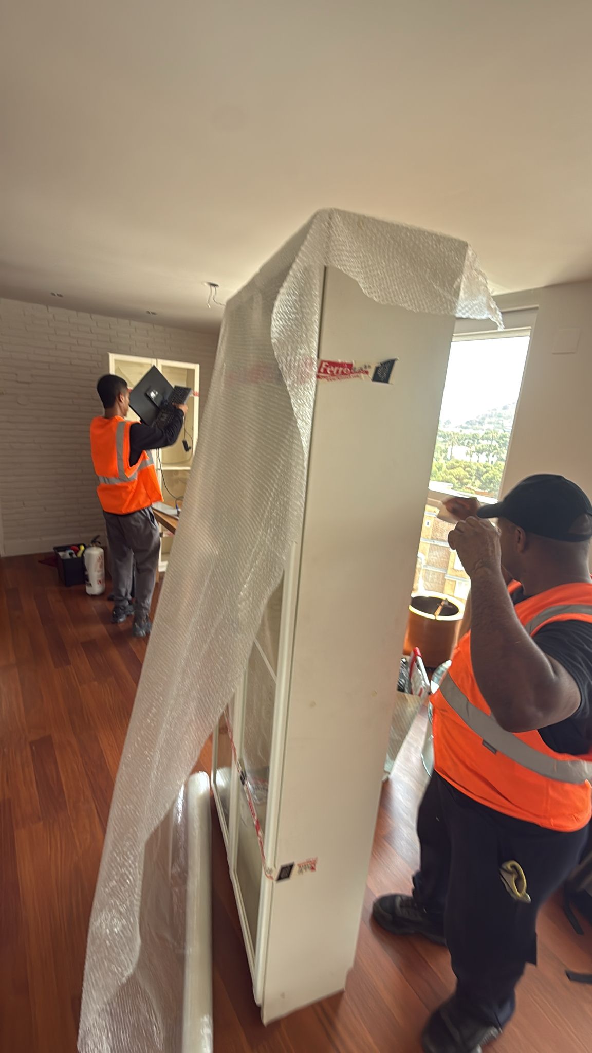 Dos trabajadores trasladando un armario alto envuelto en plástico de burbujas